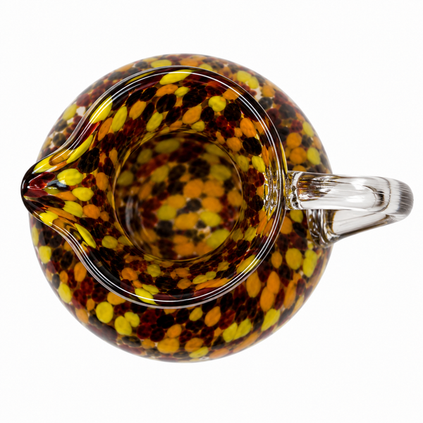 A clear glass pitcher with a rounded body, small spout, and curved handle, decorated with a dense speckled pattern of yellow, orange, and dark brown bubbles on a transparent base, shown from a top-down angle on a white background.